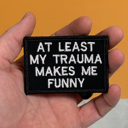 A hand holding a rectangular black embroidered patch with white text that reads 'AT LEAST MY TRAUMA MAKES ME FUNNY' against a solid orange background.