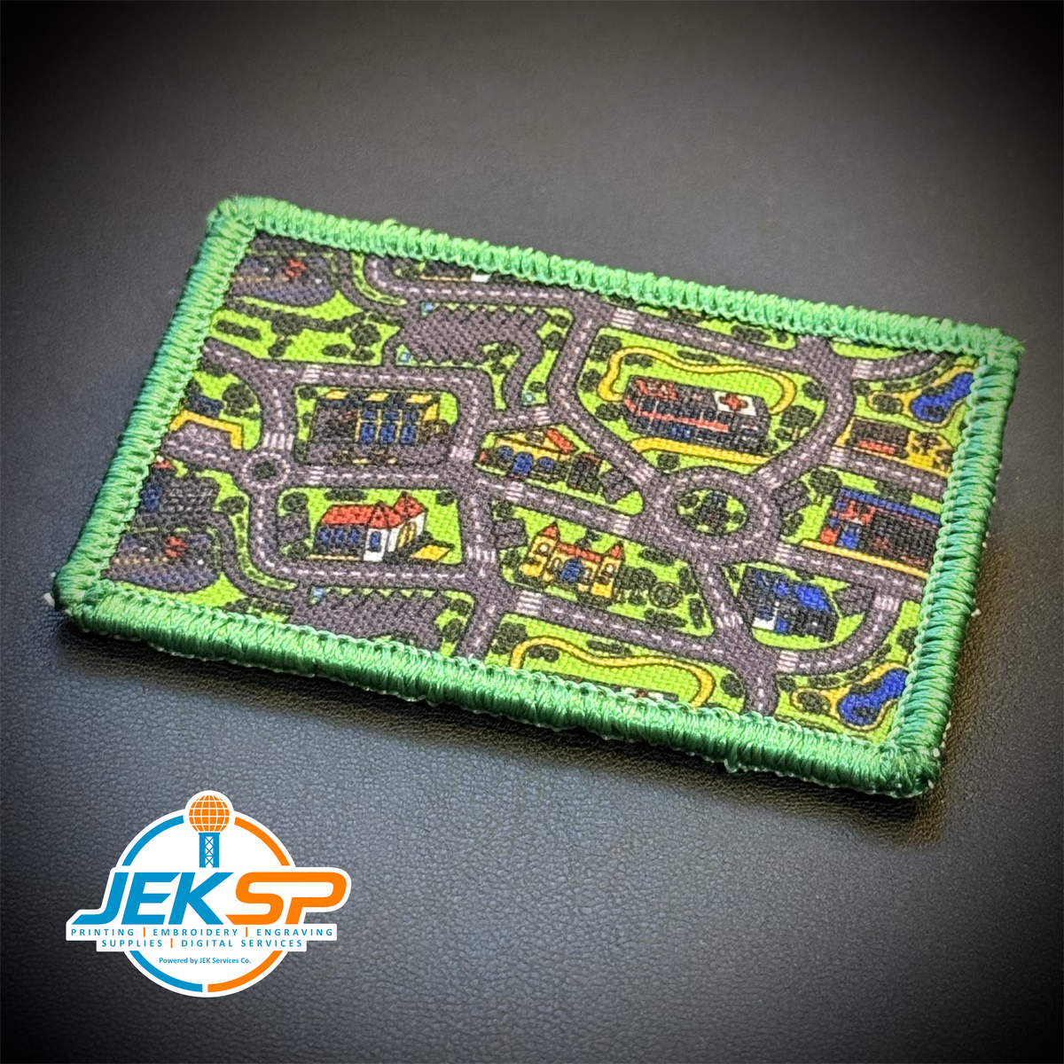A rectangular embroidered patch featuring a detailed street map design with roads, houses, trees, and blue cars, finished with a green stitched border, shown on a dark textured background with a JEKSP logo in the lower-left corner.