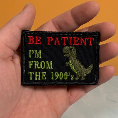 A hand holding a rectangular embroidered patch that reads 'BE PATIENT I'M FROM THE 1900's' with a green dinosaur silhouette on a black background.