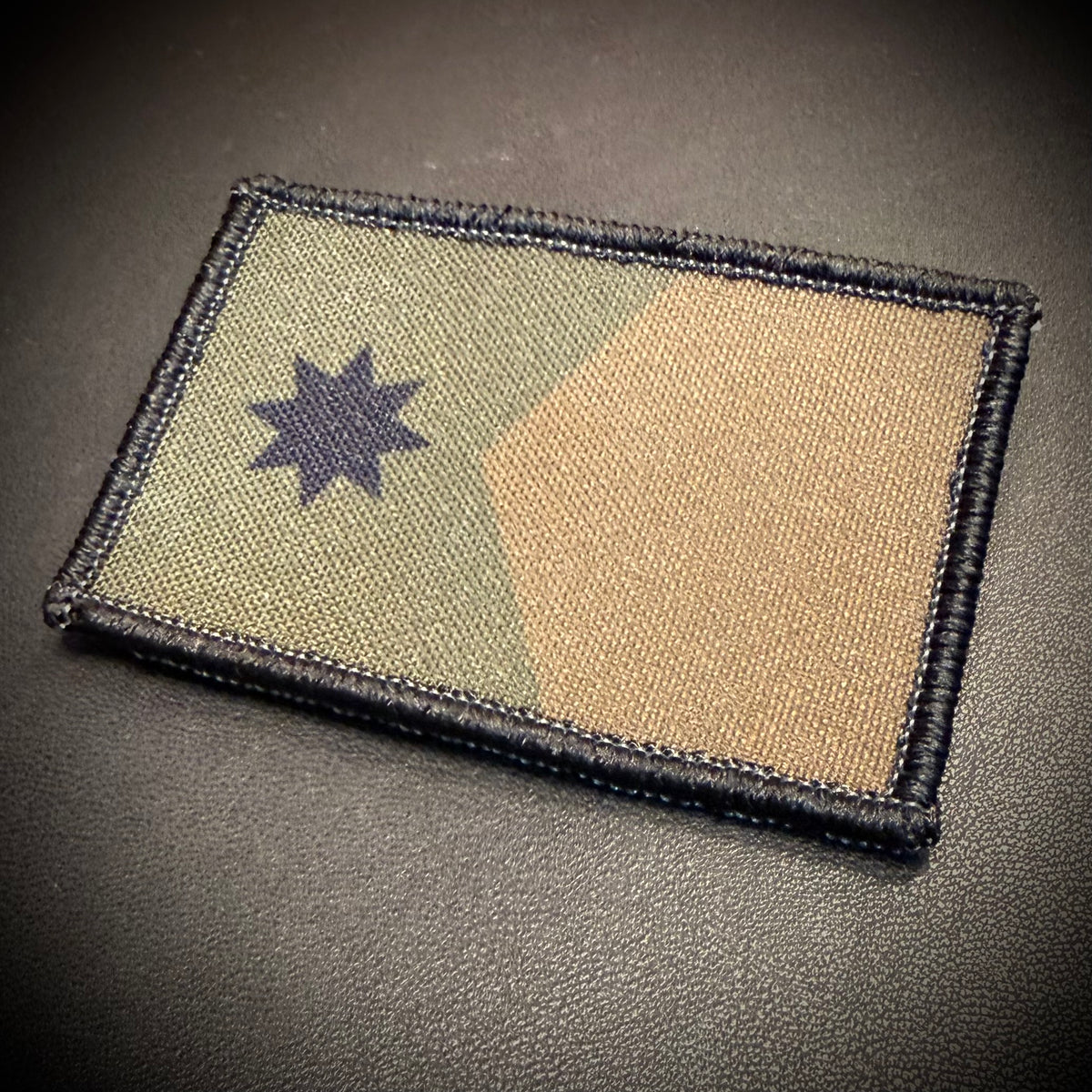 A rectangular embroidered tactical patch featuring a split camouflage design of olive green and tan with a dark navy blue eight-pointed star, shown on a dark textured background.