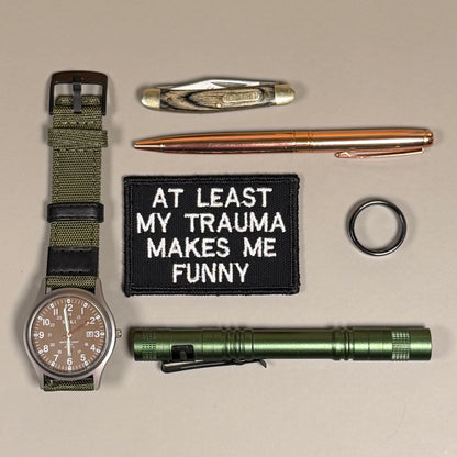 A flat-lay arrangement of tactical and utility accessories on a beige background, including a military-style watch with an olive green fabric strap, a copper-colored pen, a black embroidered patch reading 'AT LEAST MY TRAUMA MAKES ME FUNNY', a green flashlight, a folding pocket knife, and a black ring.