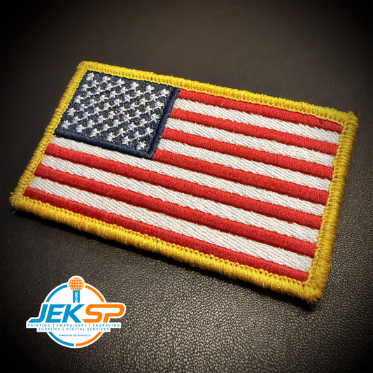 An embroidered United States flag patch with a yellow border, displayed on a dark textured surface. A 'JEK SP' brand logo is visible in the bottom-left corner.