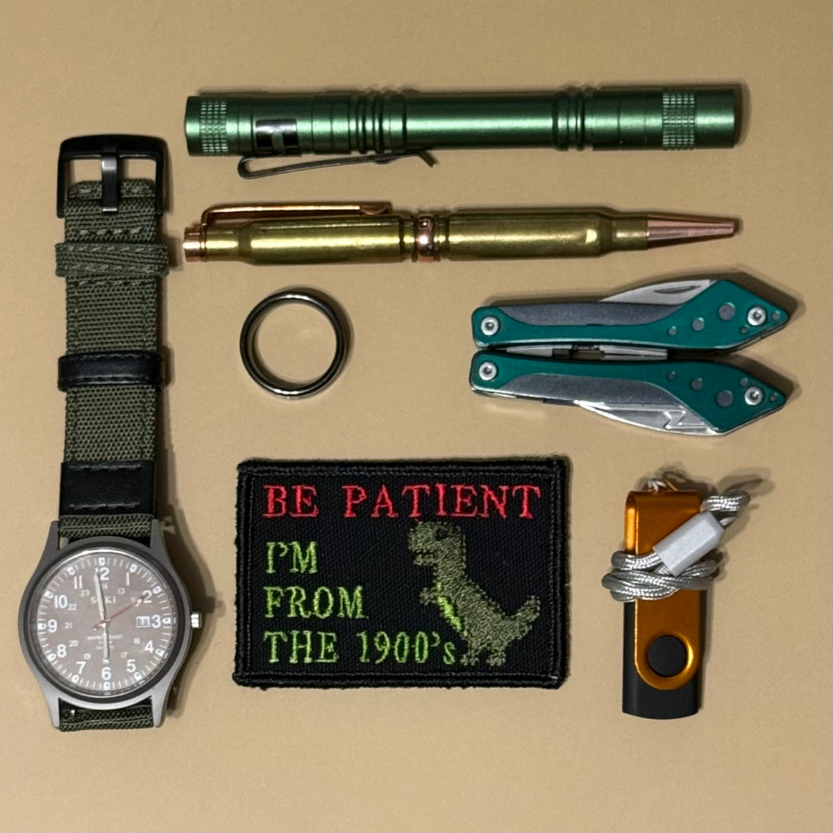 A flat-lay collection of tactical and utility items on a tan background, including a watch with an olive green fabric strap, a green cylindrical flashlight, a brass-toned pen, a folding pocket knife, a green folding multi-tool, a black embroidered patch reading 'BE PATIENT I'M FROM THE 1900's' with a dinosaur graphic, a black keychain ring, and an orange and black cord utility tool.