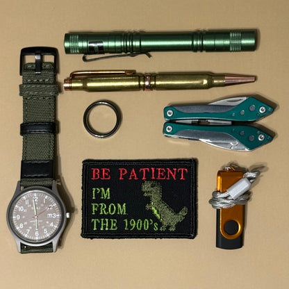 A flat-lay collection of tactical and utility items on a tan background, including a watch with an olive green fabric strap, a green cylindrical flashlight, a brass-toned pen, a folding pocket knife, a green folding multi-tool, a black embroidered patch reading 'BE PATIENT I'M FROM THE 1900's' with a dinosaur graphic, a black keychain ring, and an orange and black cord utility tool.