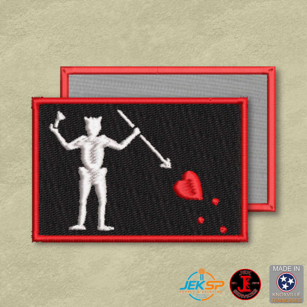 Embroidered patch with skeleton holding a heart and two daggers on a black background, with red border, on a beige background.