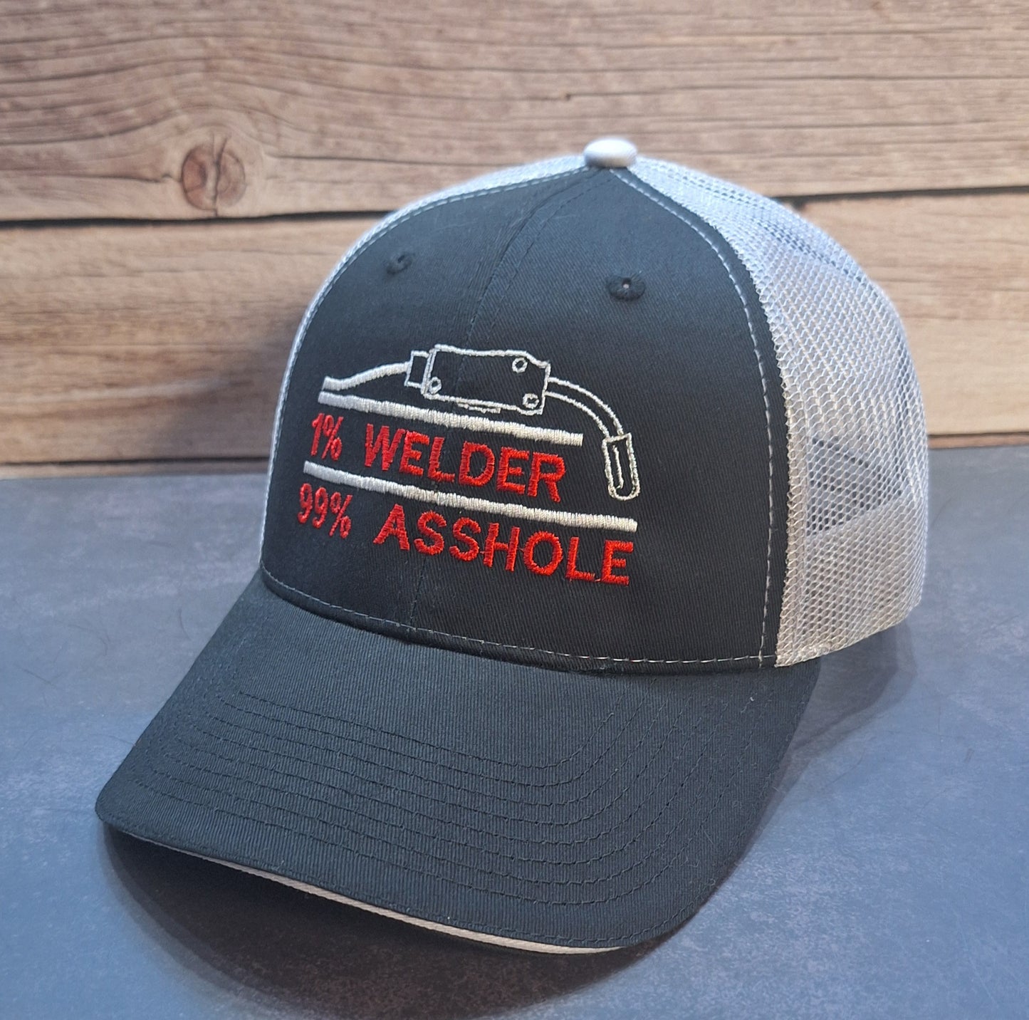 Black and white cap with '1% Welder 99% Asshole' text on a wooden surface