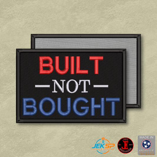 Built Not Bought embroidered morale patch with red, white, and blue lettering on black fabric, genuine hook backing, and gray loop panel displayed behind.