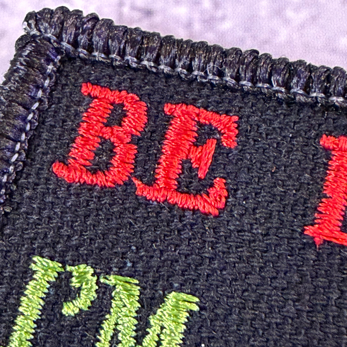 Close-up of a black embroidered fabric patch with red and green lettering on a textured background.