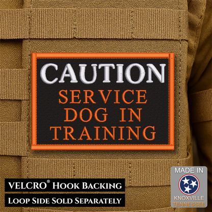 A rectangular embroidered patch with a black background and orange border, reading “CAUTION SERVICE DOG IN TRAINING,” shown attached to tan tactical gear. Text at the bottom says VELCRO™ Hook Backing and LOOP SIDE SOLD SEPARATELY, and a “Made in Knoxville Tennessee” badge appears in the lower right.