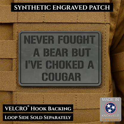 I’ve Choked A Cougar laser-engraved synthetic morale patch: a dark gray rectangular synthetic engraved patch with embossed text reading 'NEVER FOUGHT A BEAR BUT I'VE CHOKED A COUGAR,' shown attached to tan tactical vest fabric.