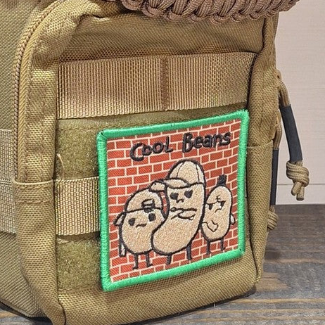 A tan tactical backpack featuring a rectangular embroidered patch with three cartoon beans on a red brick background and the text 'Cool Beans'.