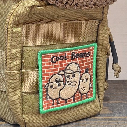 A tan tactical backpack featuring a rectangular embroidered patch with three cartoon beans on a red brick background and the text 'Cool Beans'.