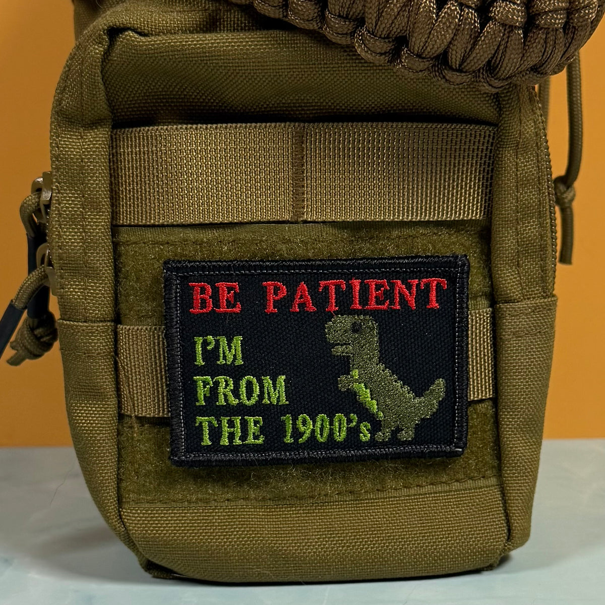 An embroidered patch on an olive green tactical pouch. The patch has a black background with red text reading 'BE PATIENT', green text reading 'I'M FROM THE 1900's', and a green dinosaur silhouette.