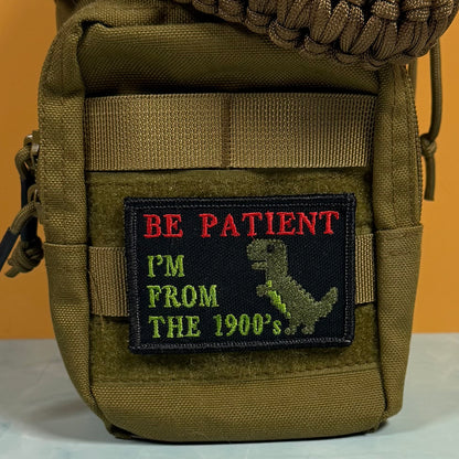 An embroidered patch on an olive green tactical pouch. The patch has a black background with red text reading 'BE PATIENT', green text reading 'I'M FROM THE 1900's', and a green dinosaur silhouette.