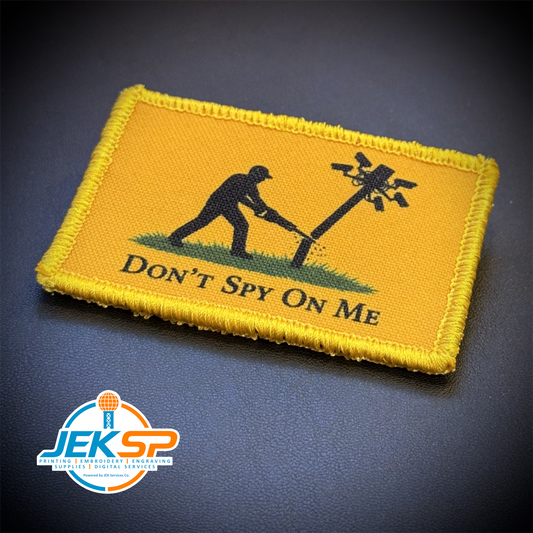 A rectangular embroidered morale patch with a yellow border and yellow field, featuring a black silhouette of a person cutting a flock camera pole with the text 'DON'T SPY ON ME'. The patch is photographed on a dark textured background with a 'JEKSP' brand watermark in the bottom-left corner.
