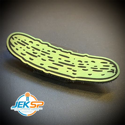 A novelty lapel pin shaped like a cucumber, featuring a light green body with black outlines and internal texture details, displayed on a dark gray background with a 'JEK SP' logo watermark in the lower-left corner.