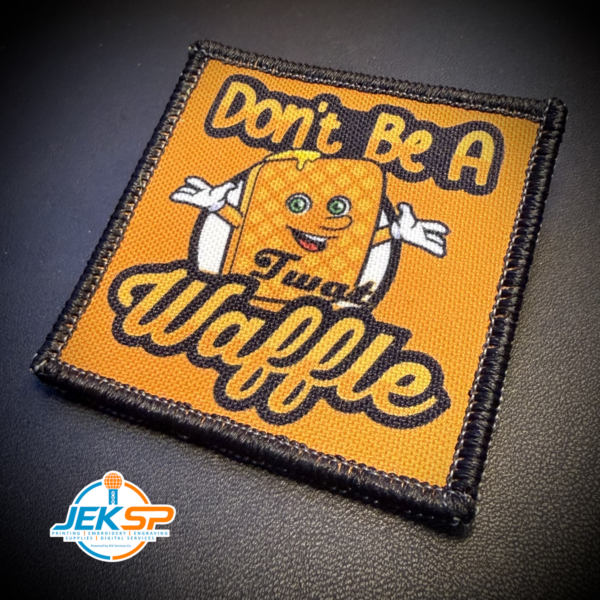 A square embroidered patch featuring a yellow-orange waffle character with the text 'Don't Be A Twat Waffle' in black and yellow lettering, shown on a dark textured background. A 'JEK SP' watermark is visible in the bottom-left corner.