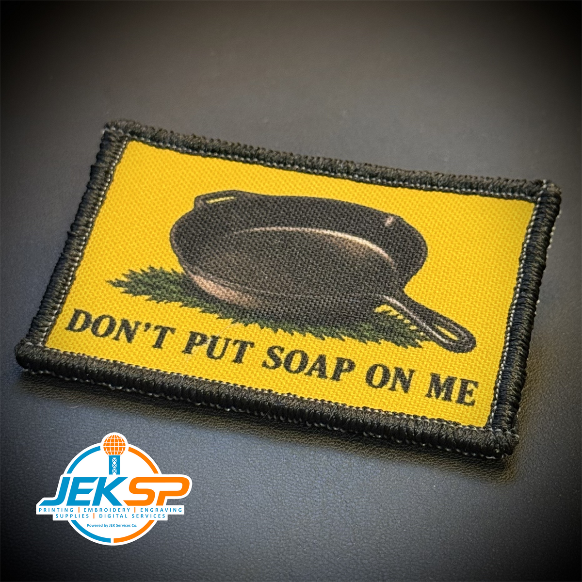 A rectangular embroidered patch with a yellow background, black border, a black cast-iron skillet, and the text 'DON'T PUT SOAP ON ME' on a dark textured surface. A 'JEKSP' watermark is visible in the bottom-left corner.