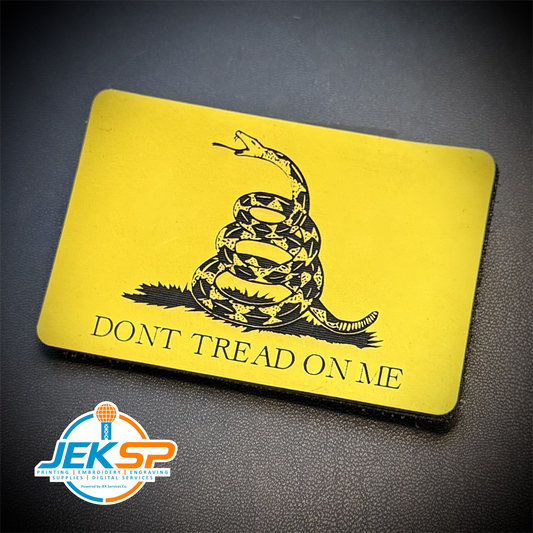 A rectangular yellow Gadsden flag-style patch or sticker with a black coiled rattlesnake graphic and the text 'DONT TREAD ON ME,' photographed on a dark textured surface. A JEKSP watermark appears in the lower-left corner.