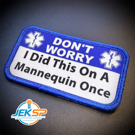A rectangular embroidered patch with a blue border and a white center, reading 'DON'T WORRY I Did This On A Mannequin Once' with two white medical-style star symbols. The patch is photographed on a dark textured surface, with a JEKSP watermark in the lower-left corner.