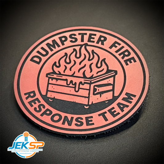 A circular rubber patch featuring a black dumpster fire graphic with flames and the text 'DUMPSTER FIRE RESPONSE TEAM' in black on a muted red background, shown on a dark textured surface.