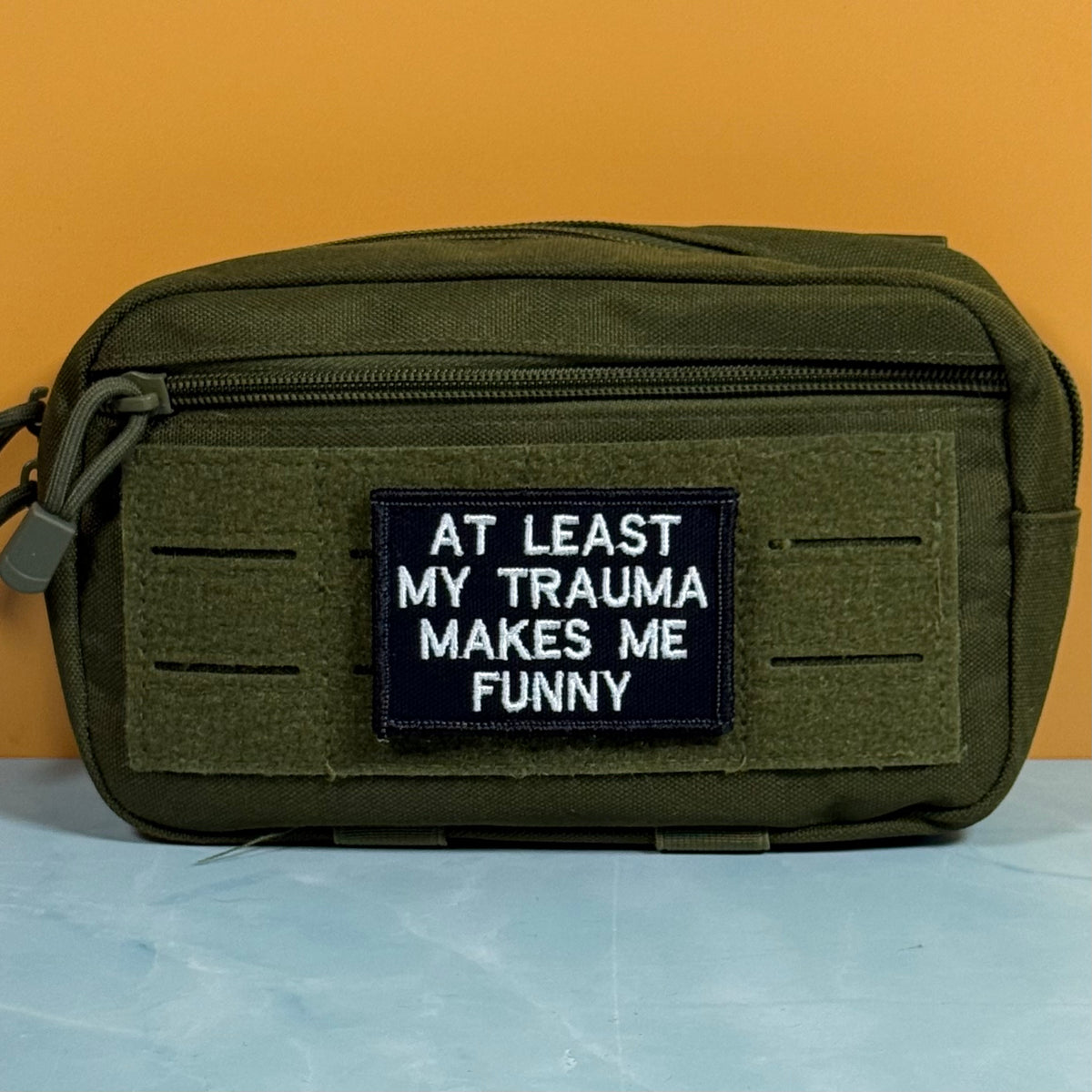 An olive green tactical pouch with a front zipper and a black rectangular patch that reads 'AT LEAST MY TRAUMA MAKES ME FUNNY' in white text, set against a solid orange background and light blue surface.