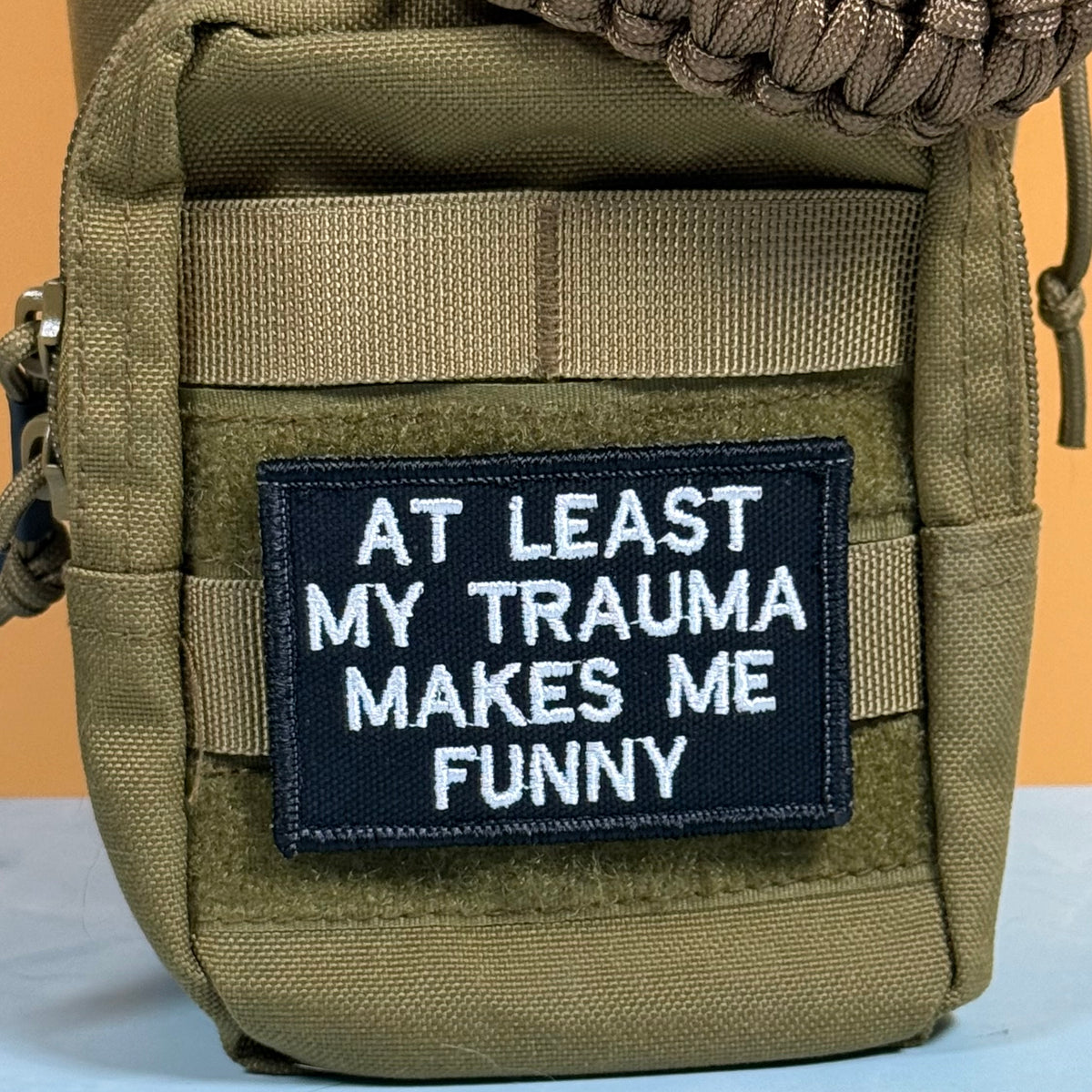 An olive green tactical pouch with a black rectangular patch that reads 'AT LEAST MY TRAUMA MAKES ME FUNNY' in white embroidered text.