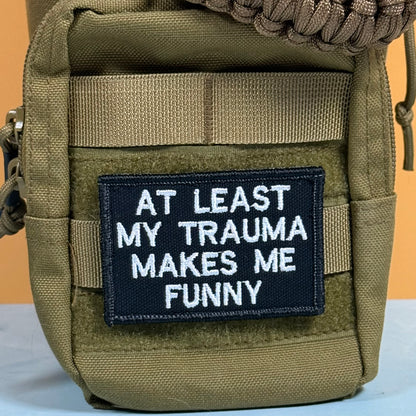An olive green tactical pouch with a black rectangular patch that reads 'AT LEAST MY TRAUMA MAKES ME FUNNY' in white embroidered text.