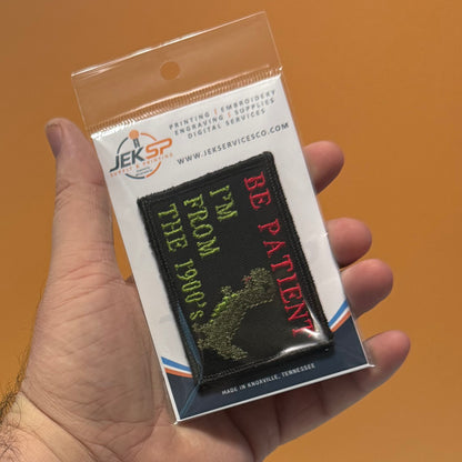 A hand holding a packaged black embroidered patch that reads 'BE PATIENT' in red and 'I'M FROM THE 1900's I'M FROM THE 1900's' in green, with a white backing card and clear plastic sleeve. The background is a solid orange surface.