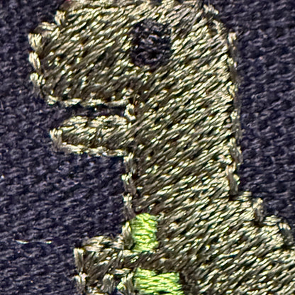 Close-up of an embroidered dinosaur motif on dark navy fabric, with a light olive-green body and a bright neon green accent.