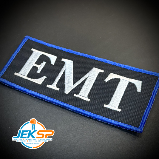 An embroidered rectangular patch with the letters 'EMT' in white on a black background, bordered in blue, photographed on a dark surface. A JEKSP watermark appears in the lower-left corner.
