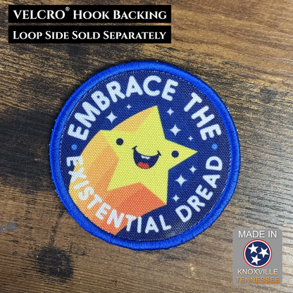 Patch with a yellow star and text 'Embrace the Existential Dread' on a wooden surface, with branding information.