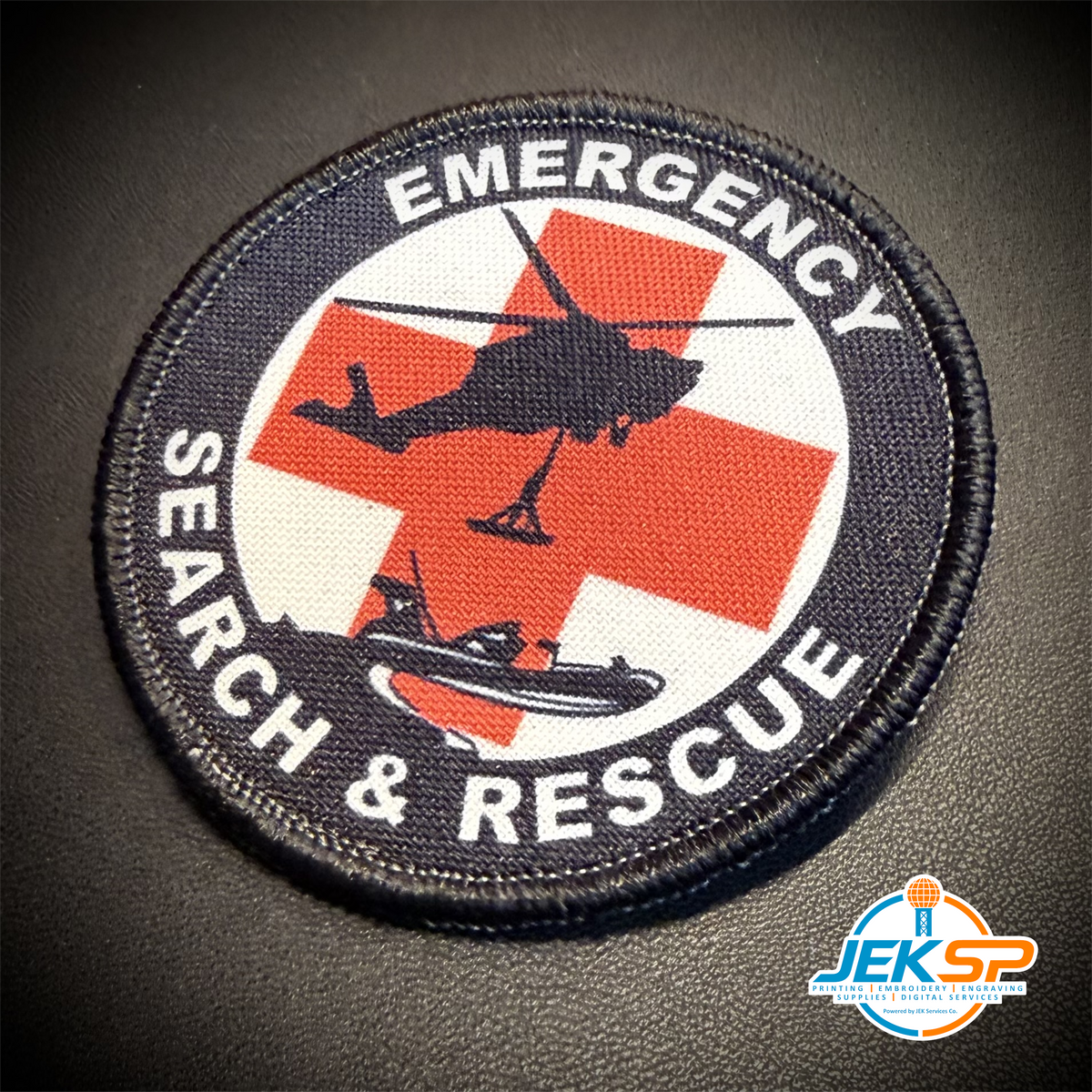 A circular embroidered patch with a black border and the words 'EMERGENCY SEARCH & RESCUE' around a red cross, helicopter, and boat graphic on a white background, shown on a dark textured surface.