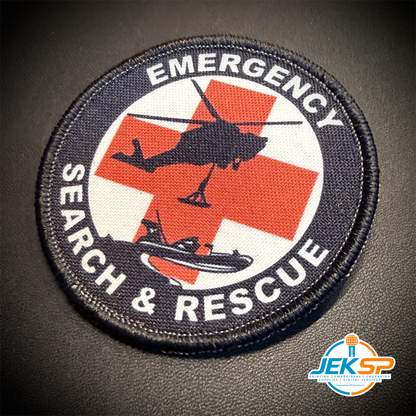 A circular embroidered patch with a black border and the words 'EMERGENCY SEARCH & RESCUE' around a red cross, helicopter, and boat graphic on a white background, shown on a dark textured surface.