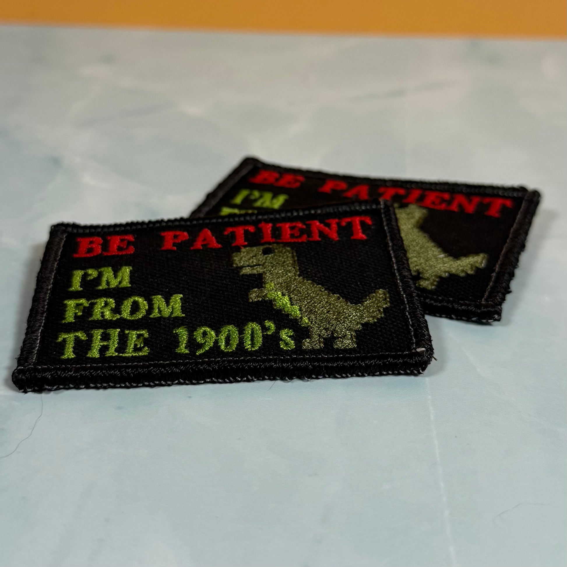 Two embroidered rectangular patches on a light marble-like surface. The front patch is black with red text reading 'BE PATIENT', lime green text reading 'I'M FROM THE 1900's', and a green dinosaur silhouette.