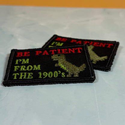 Two embroidered rectangular patches on a light marble-like surface. The front patch is black with red text reading 'BE PATIENT', lime green text reading 'I'M FROM THE 1900's', and a green dinosaur silhouette.