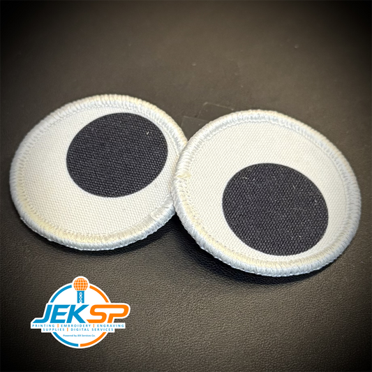 Two round white fabric patches with black circular centers, resembling cartoon eyes, shown on a dark textured background. A JEKSP logo is visible in the bottom-left corner.