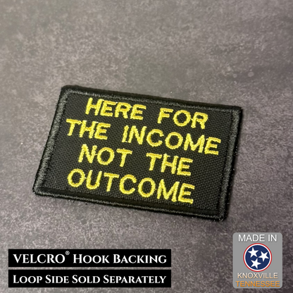Black patch with yellow text 'HERE FOR THE INCOME NOT THE OUTCOME' on a gray background, with VELCRO hook backing information.