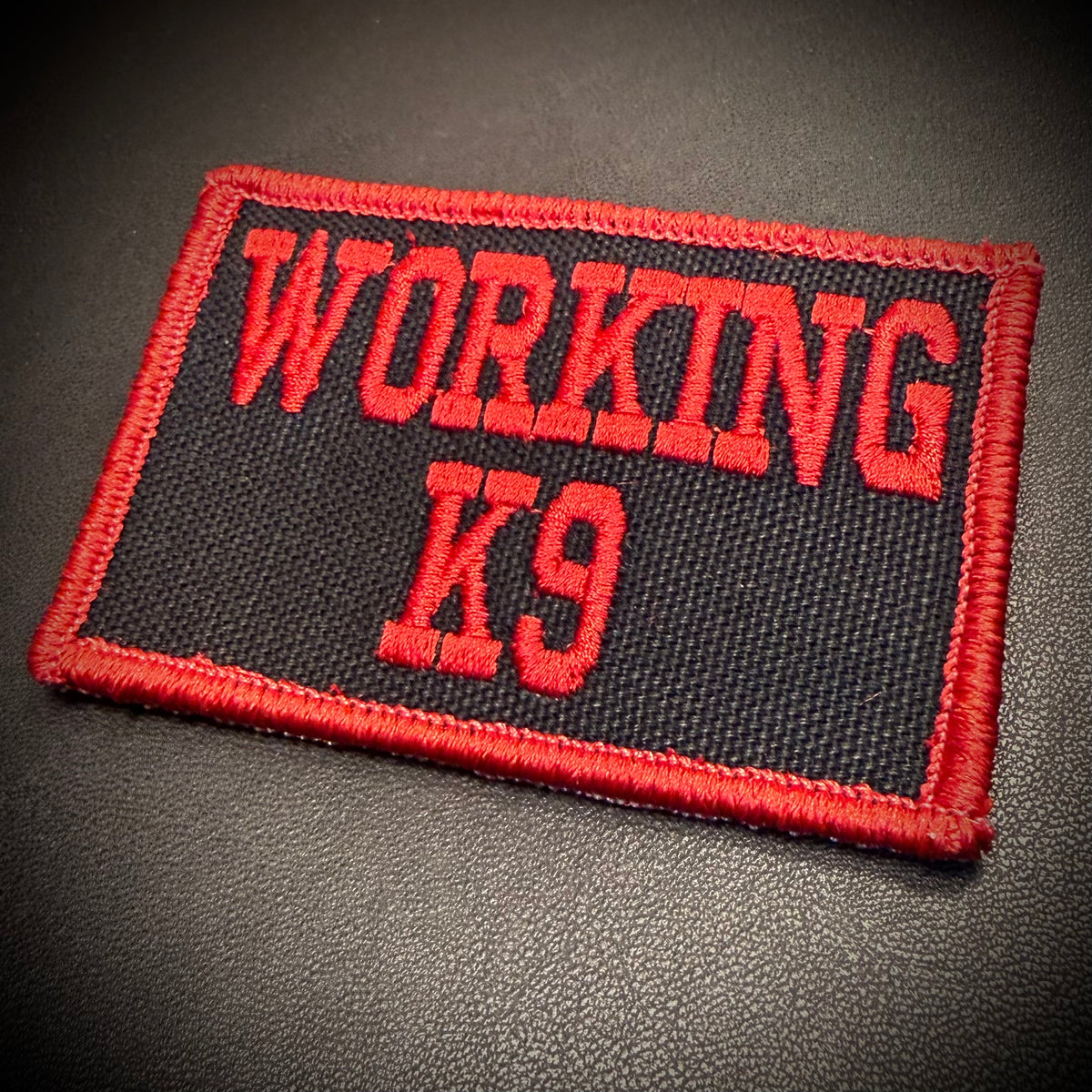 A rectangular embroidered patch with the words 'WORKING K9' in red lettering on a black background, finished with a red stitched border, shown on a dark textured surface.