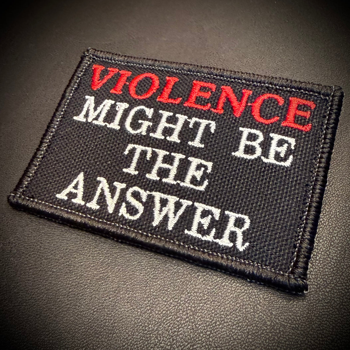 A rectangular embroidered patch with the text 'VIOLENCE MIGHT BE THE ANSWER' in red and white lettering on a black background, photographed on a dark textured surface.