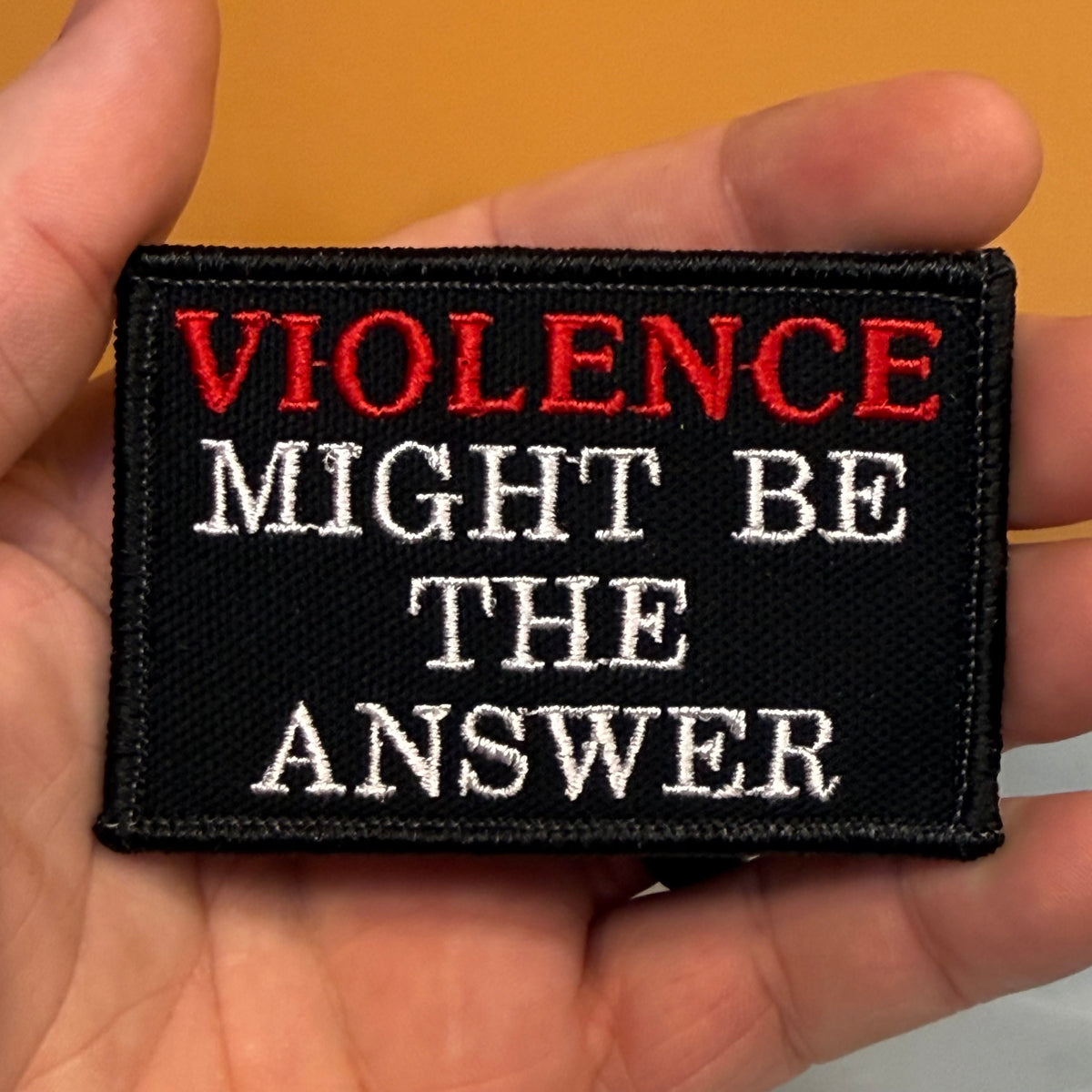 A rectangular black embroidered patch held in a hand, featuring the text 'VIOLENCE MIGHT BE THE ANSWER' in red and white lettering, against a warm tan background.