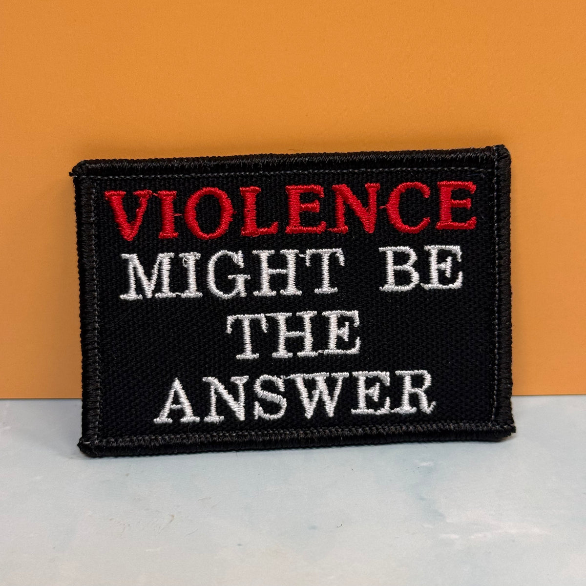 A rectangular embroidered patch with the text 'VIOLENCE MIGHT BE THE ANSWER' in red and white lettering on a black background, placed against an orange wall and a light surface.