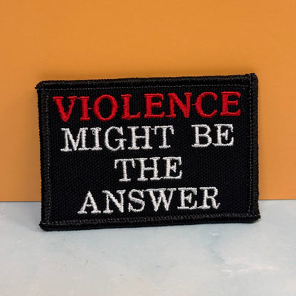 A rectangular embroidered patch with the text 'VIOLENCE MIGHT BE THE ANSWER' in red and white lettering on a black background, placed against an orange wall and a light surface.