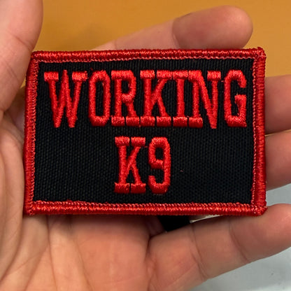 A rectangular embroidered patch held in a hand, featuring the words 'WORKING K9' in red embroidery on a black background with a red stitched border.