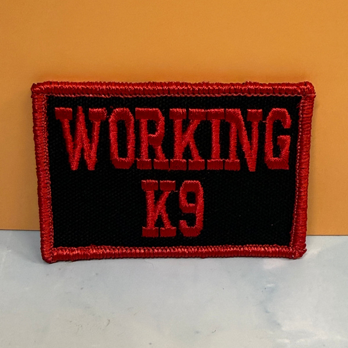 An embroidered rectangular patch with the words 'WORKING K9' in red lettering on a black background, finished with a red stitched border, photographed against a tan wall and light surface.