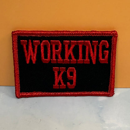 An embroidered rectangular patch with the words 'WORKING K9' in red lettering on a black background, finished with a red stitched border, photographed against a tan wall and light surface.