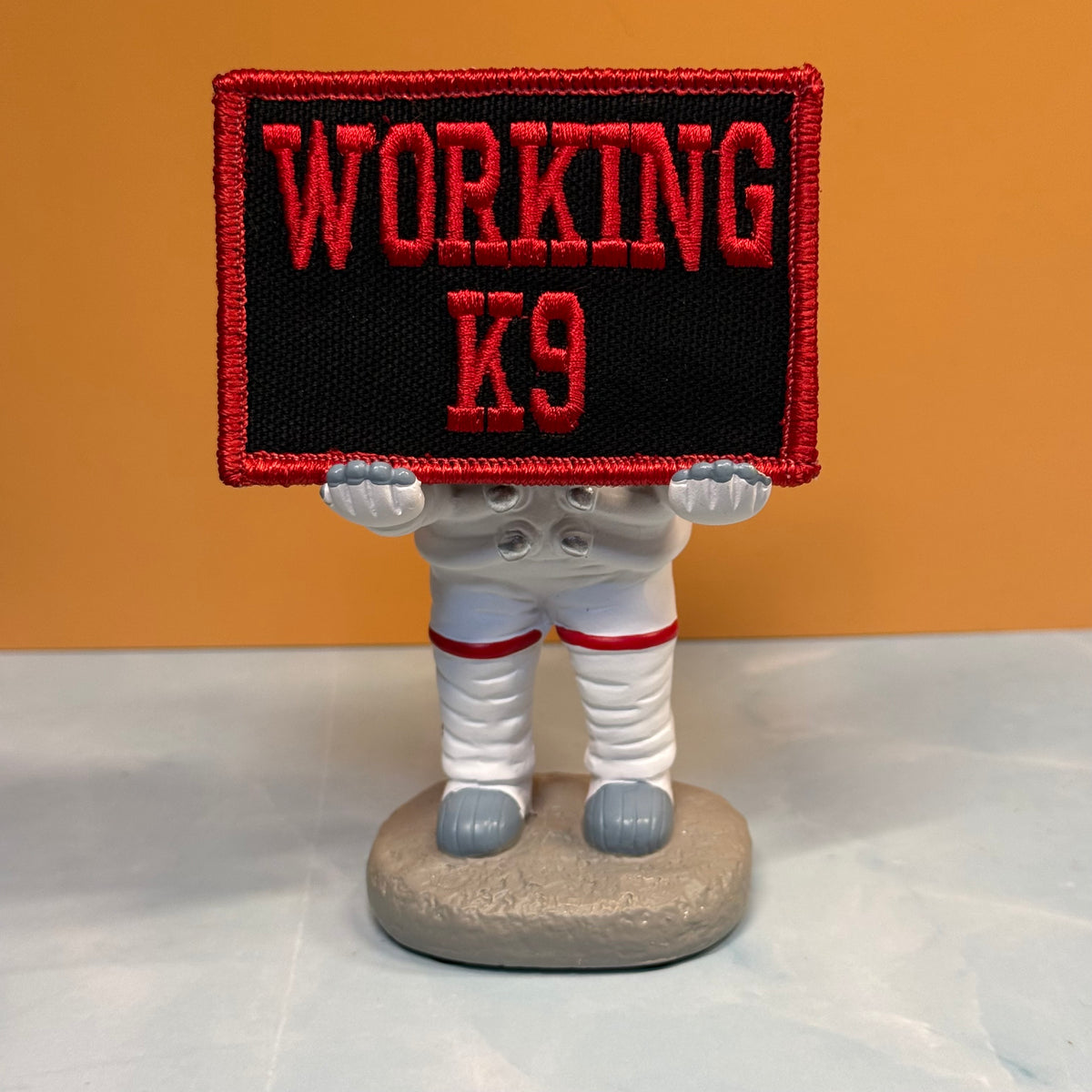 A small astronaut figurine holding a rectangular embroidered patch that reads “WORKING K9” in red on black with a red border, set against a warm tan background and light gray surface.