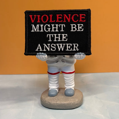 A novelty figurine of an astronaut holding a black sign that reads 'VIOLENCE MIGHT BE THE ANSWER' in red and white embroidery, set against a solid orange background.