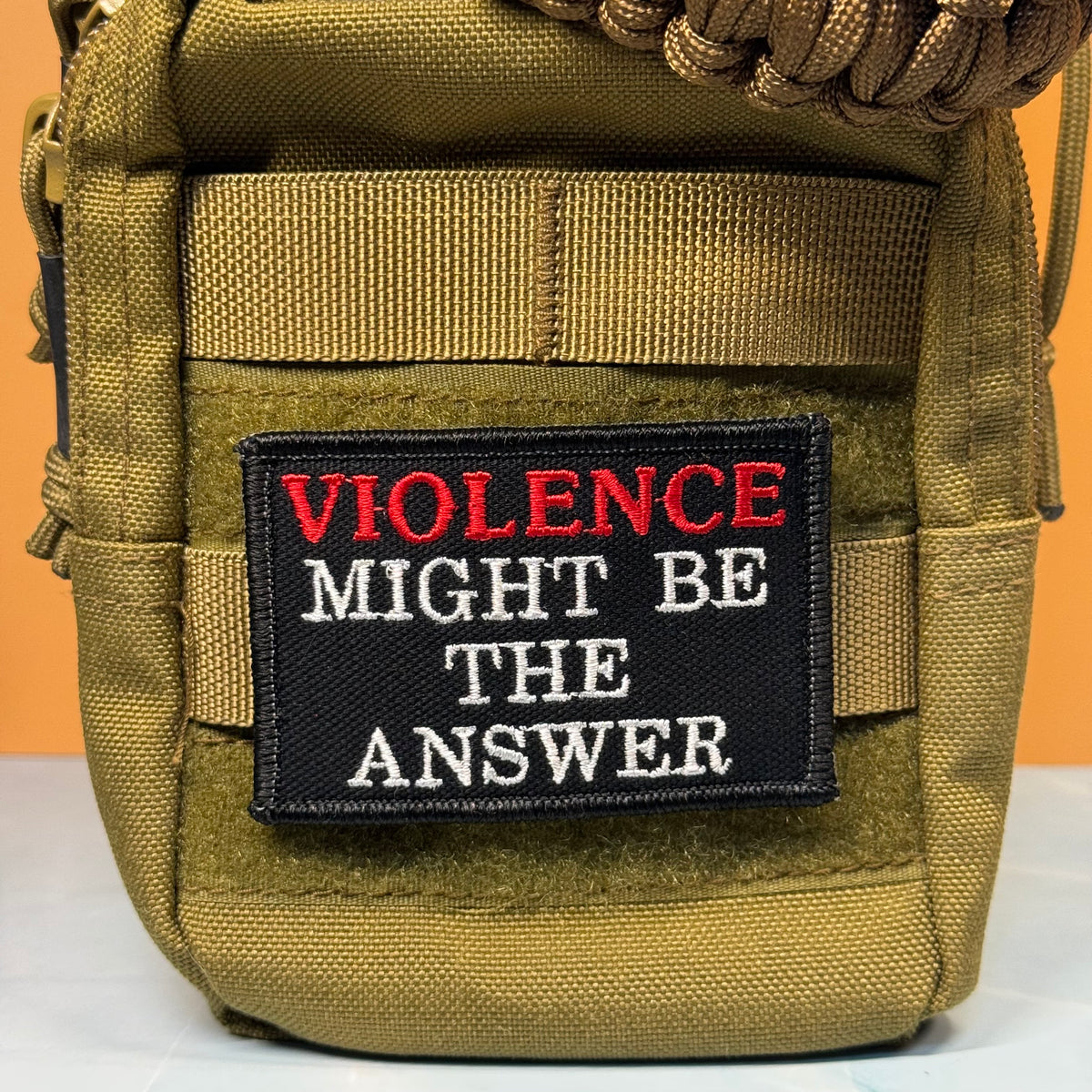 An olive drab tactical pouch with a black embroidered patch that reads 'VIOLENCE MIGHT BE THE ANSWER' in red and white lettering, set against a solid orange background.