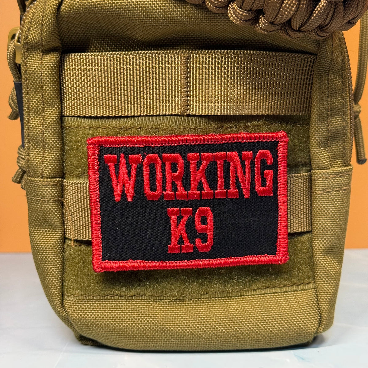 A close-up of a tactical pouch in olive drab green nylon with a black rectangular patch featuring red embroidered text that reads 'WORKING K9'.
